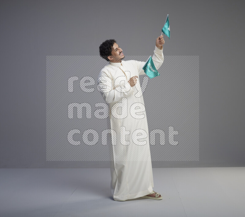 A Saudi man standing wearing thob holding small Saudi flag on gray background