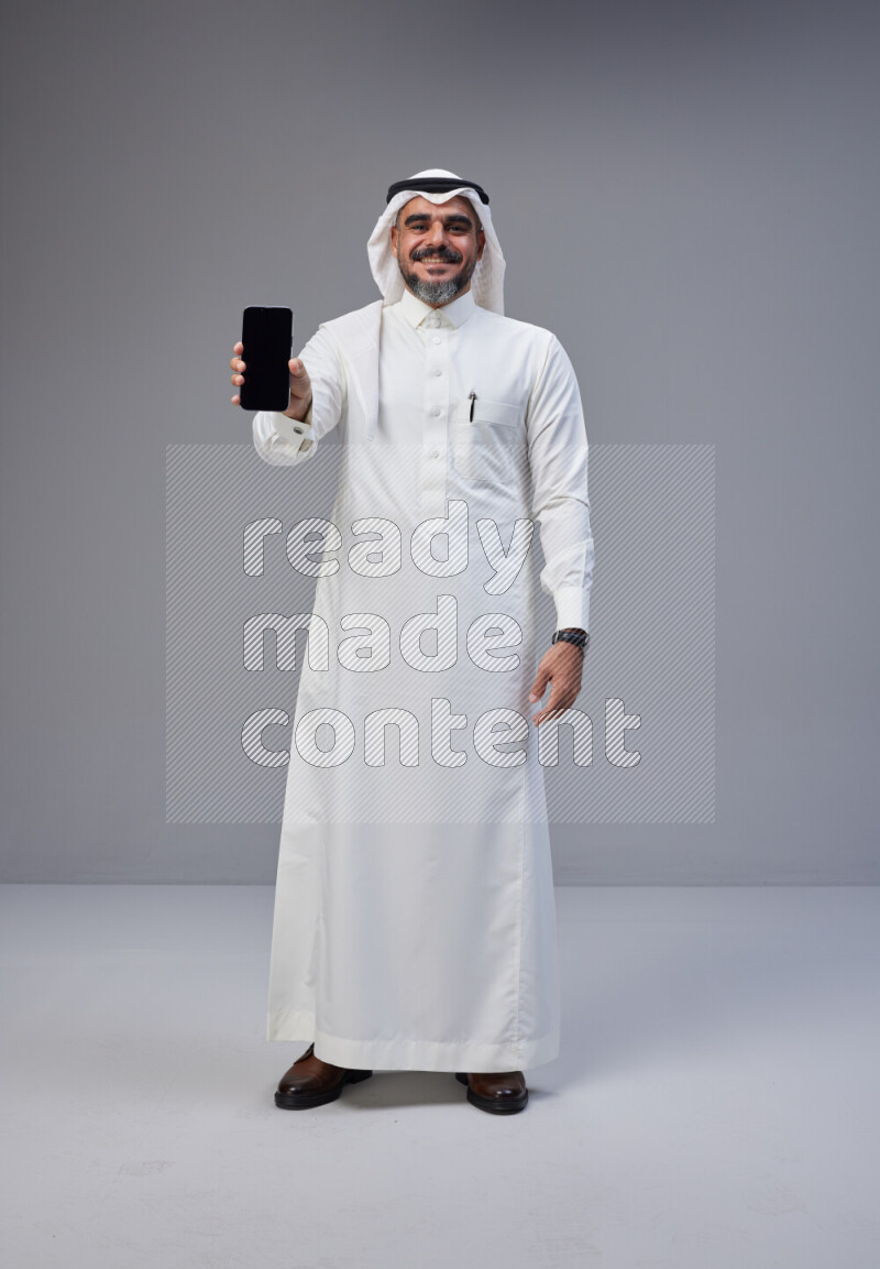 Saudi man Wearing Thob and white Shomag standing showing phone to camera on Gray background