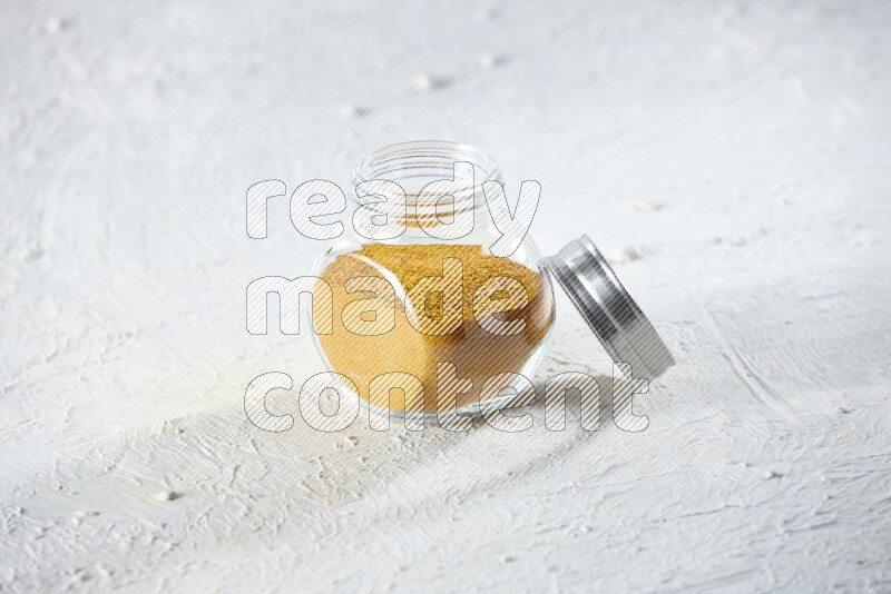 A glass spice jar full of turmeric powder on a textured white flooring