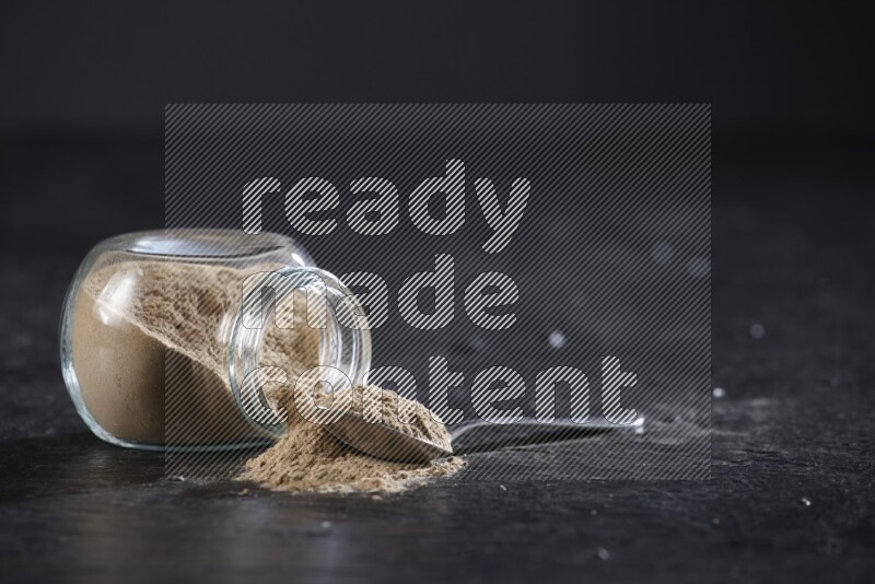 وعاء بهارات ممتلئ ببودرة الثوم مقلوب والبودرة تتناثر منه مع ملعقة معدنية مملوءة بجانبه على خلفية سوداء