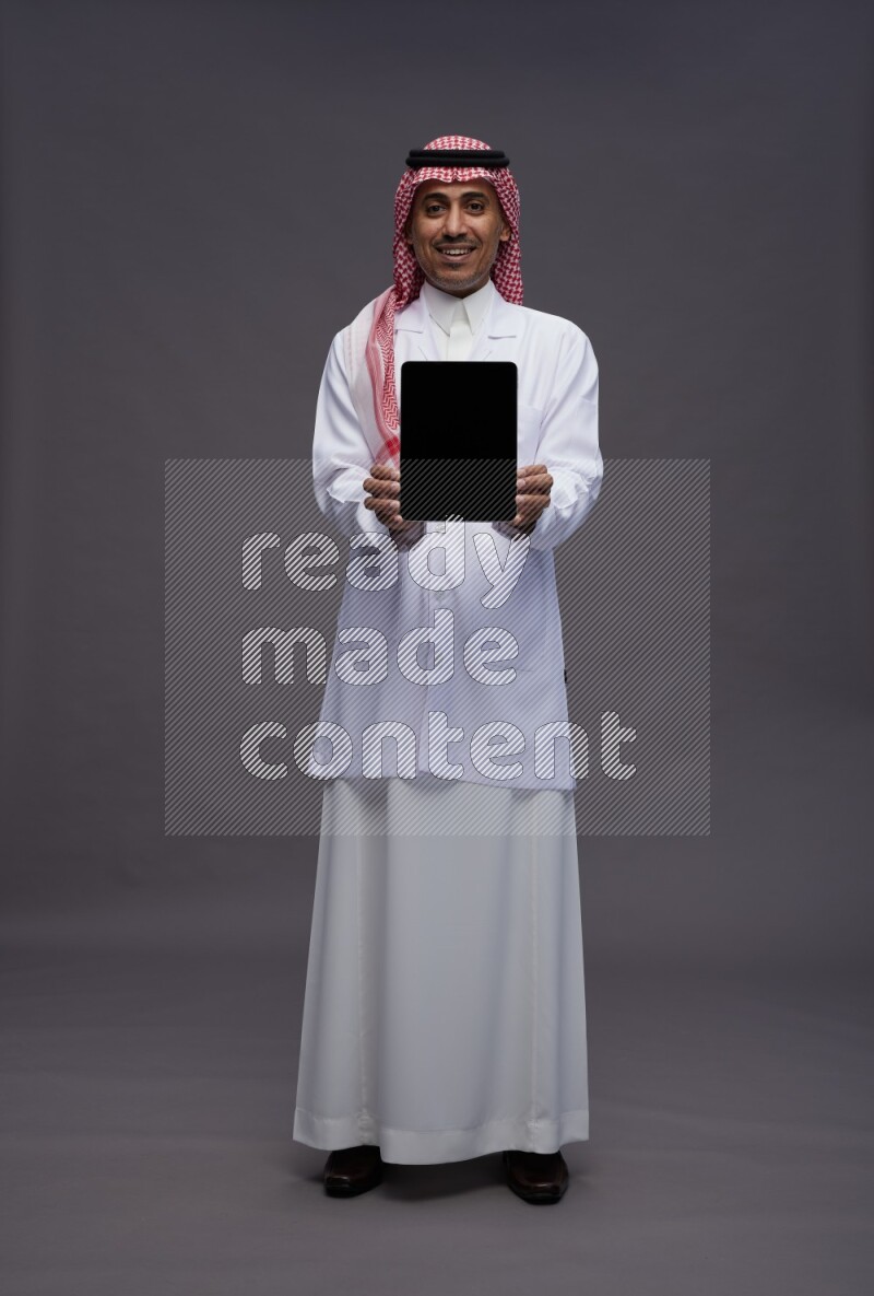 Saudi man wearing thob with lab coat and shomag with pocket employee badge standing showing tablet to camera on gray background
