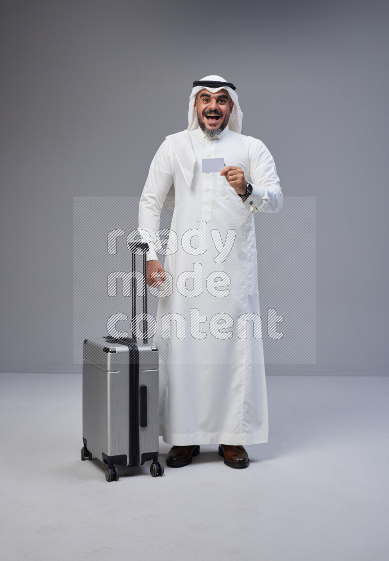 Saudi man wearing Thob and white Shomag standing holding Travel bag and ATM card on Gray background