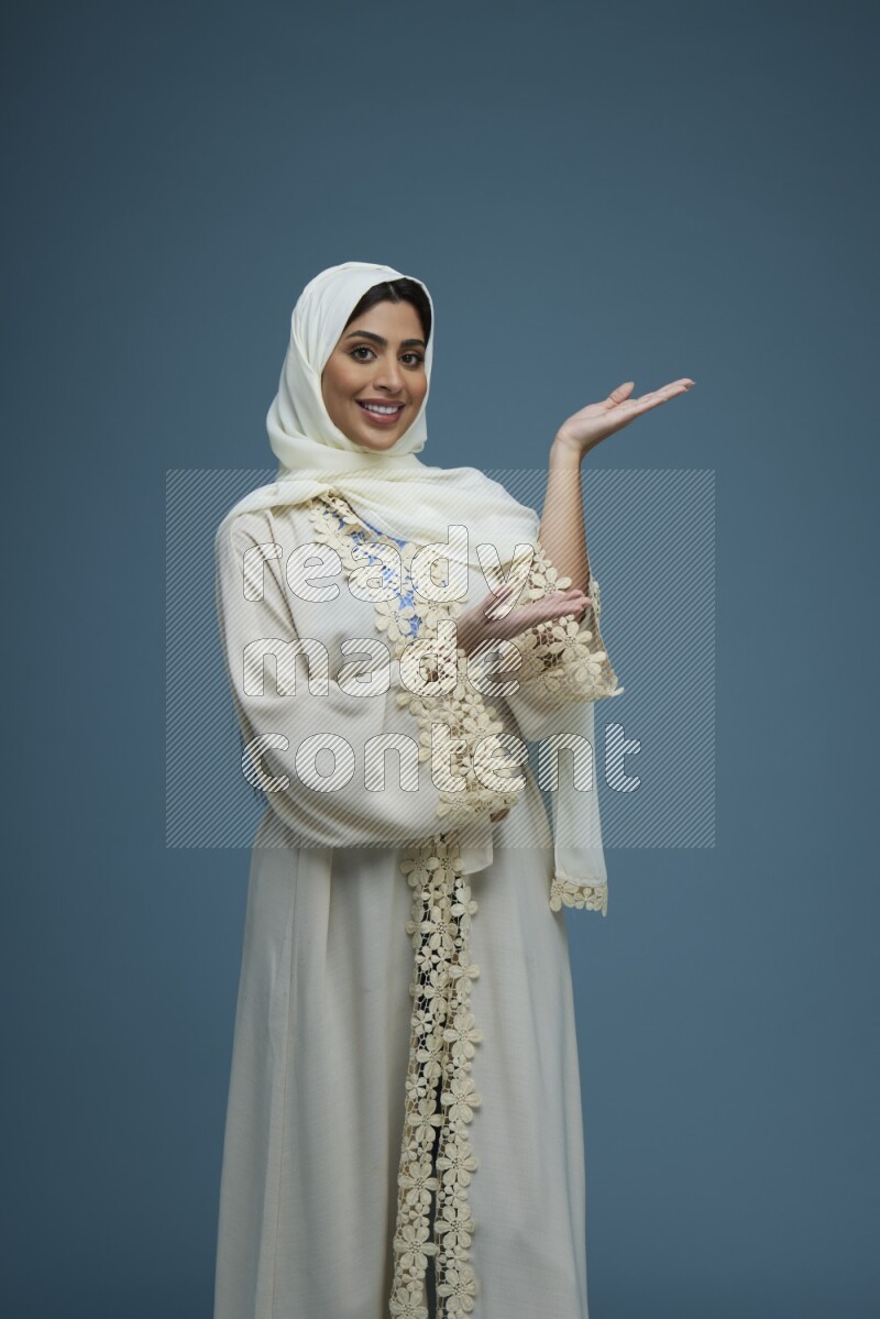 A Saudi woman pointing in a blue background wearing an off-white Abaya Hijab