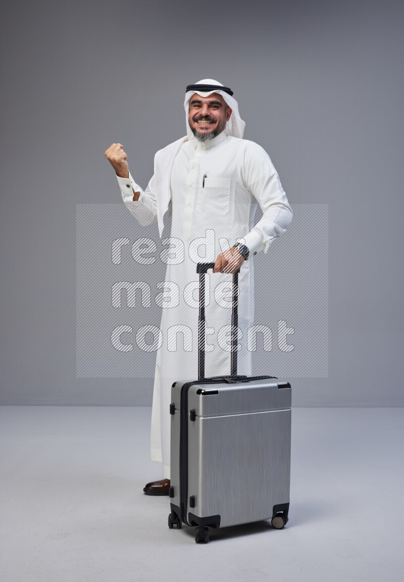 Saudi man wearing Thob and white Shomag standing holding Travel bag on Gray background