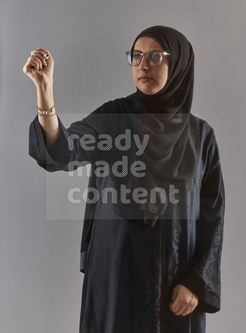 Woman in black abaya, hijab and glasses holding a marker pen to write on a transparent board with different reactions and expressions on grey background