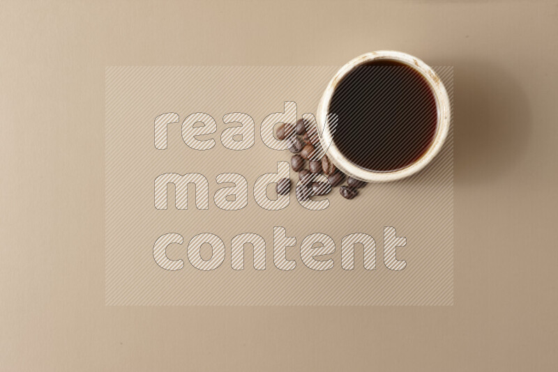 A beige pottery cup of coffee surrounded by roasted coffee beans on beige background