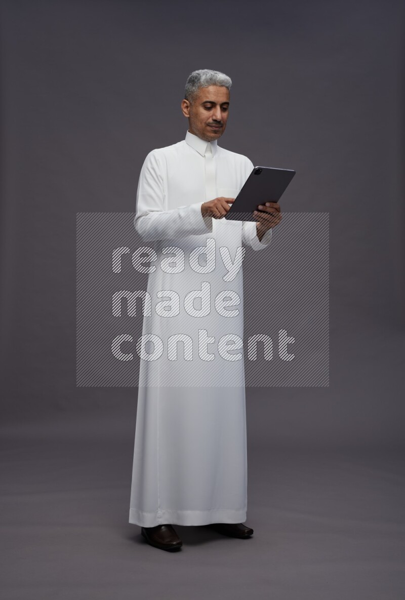Saudi man wearing thob standing working on tablet on gray background