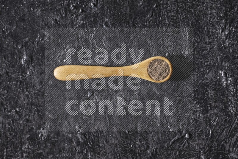 A wooden spoon full of black pepper powder on a textured black flooring