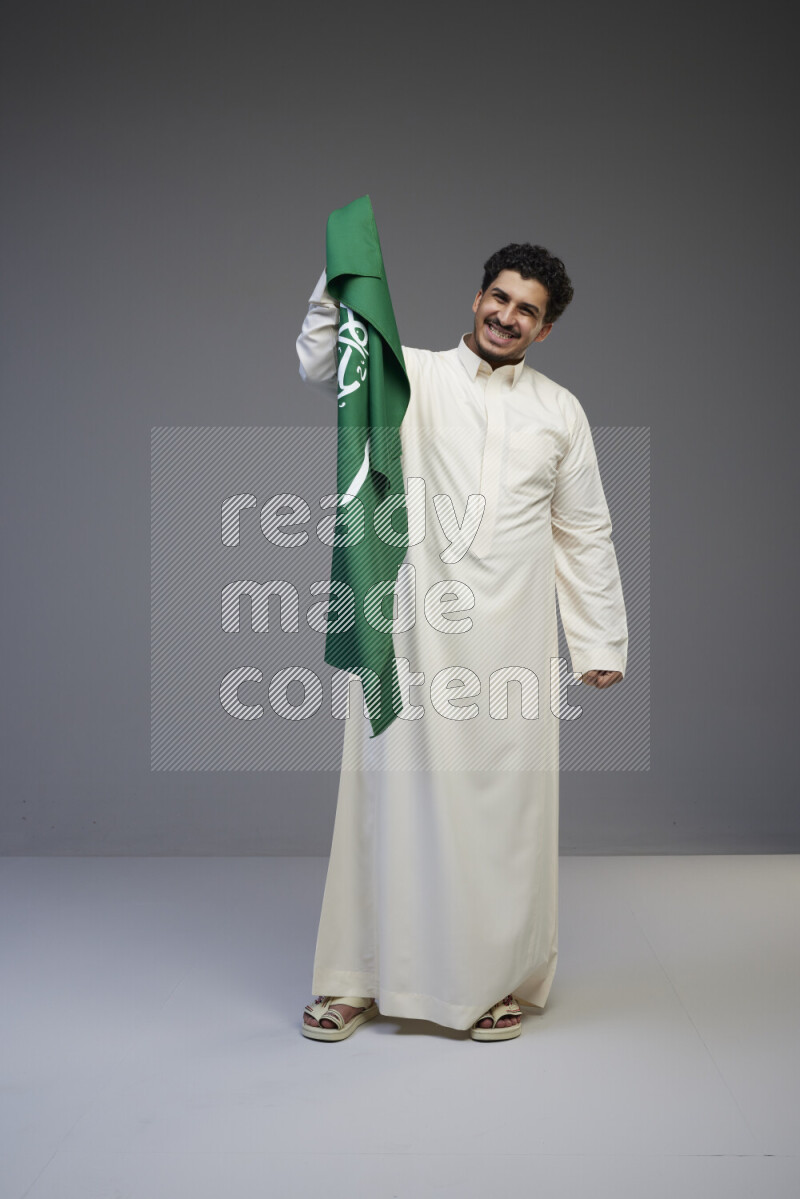 A Saudi man standing wearing thob holding big Saudi flag on gray background