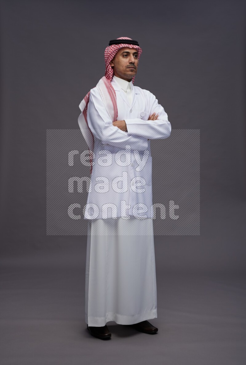 Saudi man wearing thob with lab coat and shomag with pocket employee badge standing with crossed arms on gray background