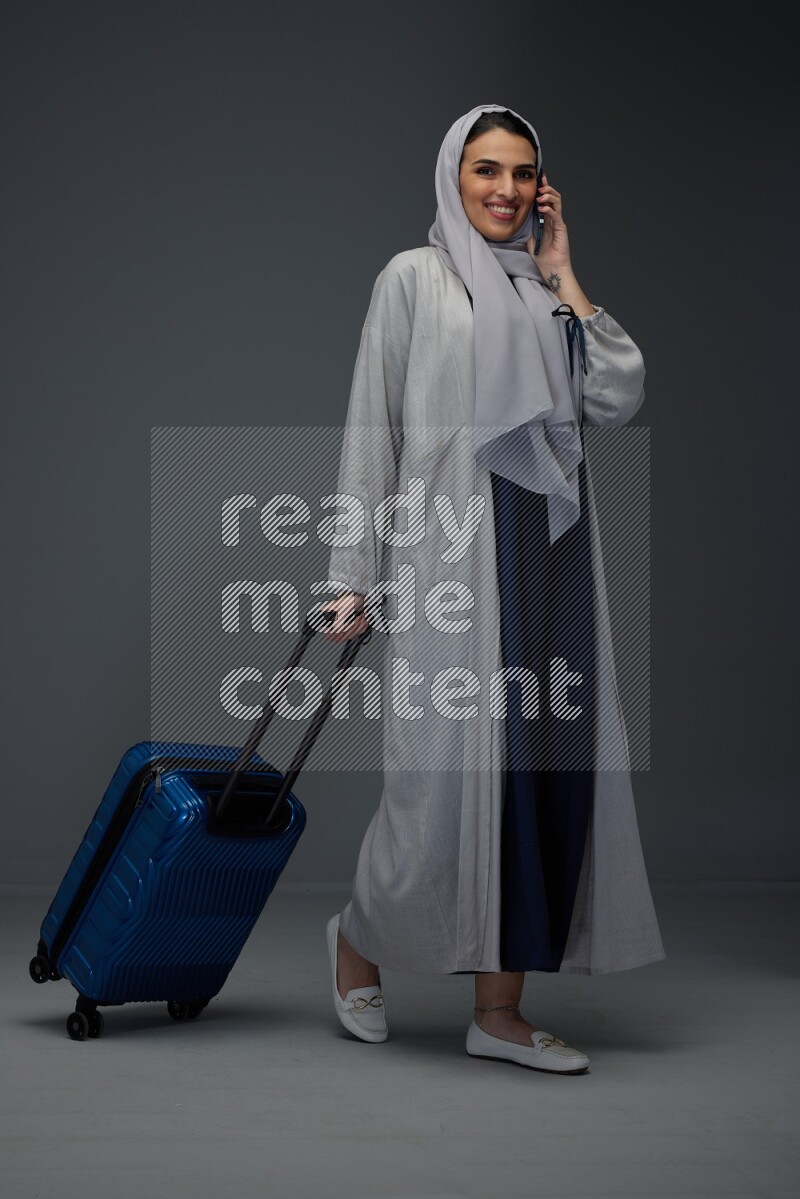 A Saudi woman wearing a light gray Abaya and head scarf standing and talking in the phone while holding shopping bags eye level on a grey background