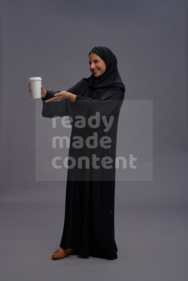 Saudi woman wearing Abaya standing holding paper cup on gray background