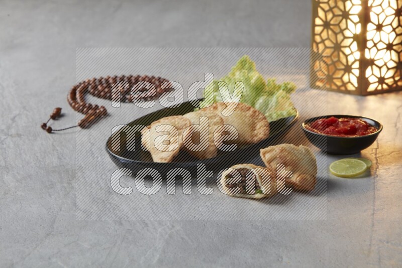 Three fried sambosas in an oval shaped black plate and a red sauce in a black round ramekin with a brown misbaha and a golden lantern on the side on a gray background