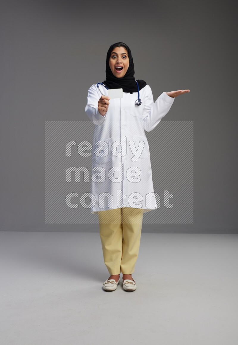 Saudi woman wearing lab coat with stethoscope standing holding ATM card on Gray background