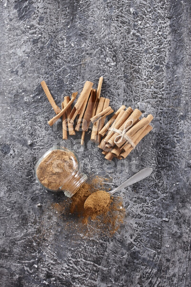 Herbal glass jar full cinnamon powder flipped and a metal spoon full of powder, cinnamon sticks stacked and bounded in the back on textured black background in different angles