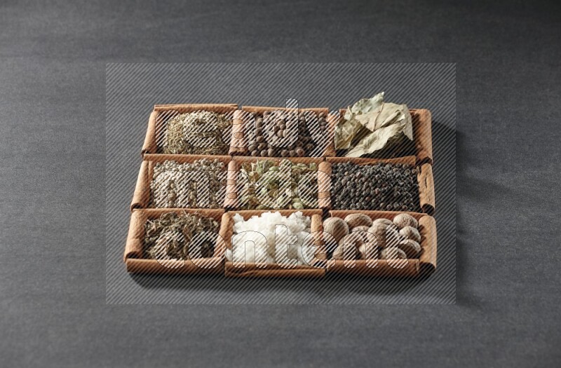 9 squares of cinnamon sticks full of cardamom in the middle surrounded by nutmeg, black pepper, bay laurel leaves, allspice, cumin, white pepper, dried basil and salt on black flooring