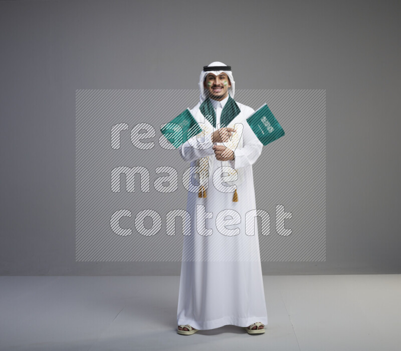 A saudi man standing wearing thob and white shomag with face painting and saudi flag scarf and holding small saudi flag on gray background