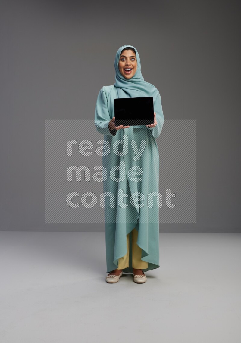Saudi Woman wearing Abaya standing showing tablet to camera on Gray background