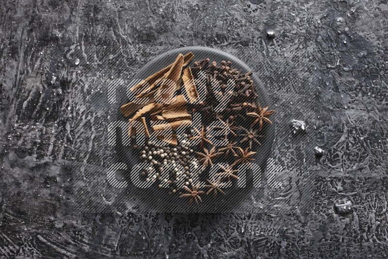 Cinnamon sticks, cloves, star anise and black and white peppers on a black plate on textured black background