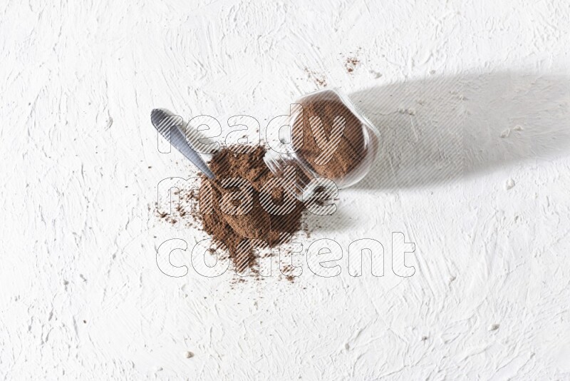 A flipped glass spice jar and a metal spoon full of cloves powder and powder came out of the jar on textured white flooring