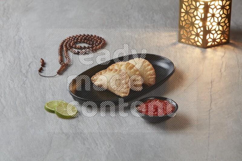Three fried sambosas in an oval shaped black plate and a red sauce in a black round ramekin with a brown misbaha and a golden lantern on the side on a gray background