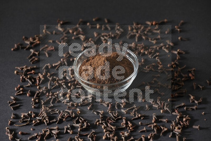 A glass bowl full of cloves powder with cloves grains spread on black flooring