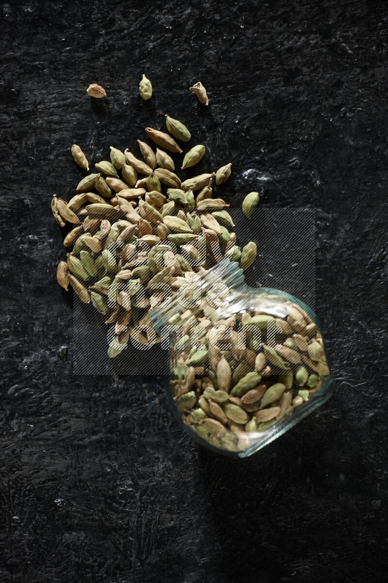 A flipped glass spice jar full of cardamom seeds on textured black flooring