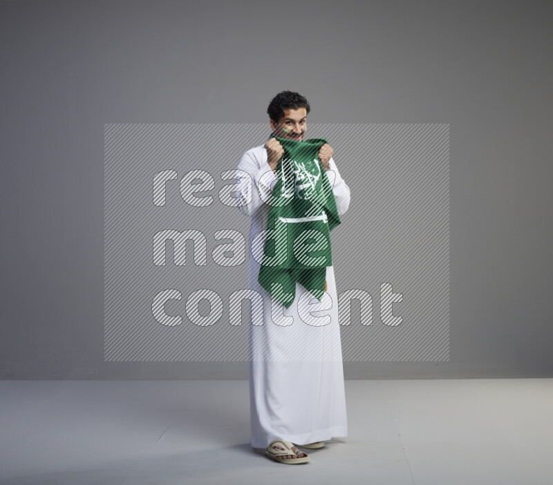 A Saudi man standing wearing thob with face painting wrapping Saudi flag scarf and holding big Saudi flag on gray background