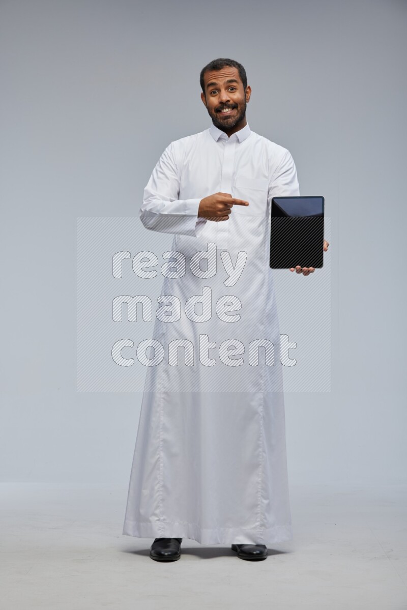 Saudi man wearing Thob standing showing tablet to camera on Gray background