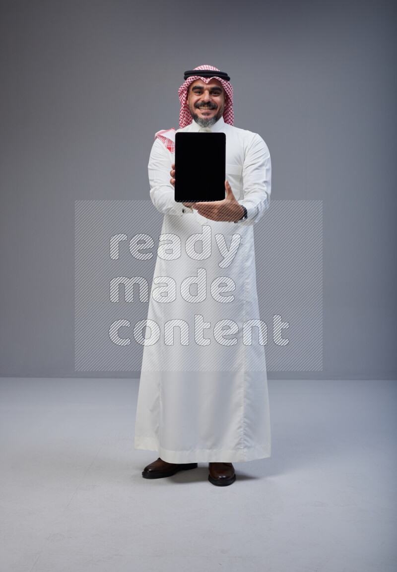 Saudi man Wearing Thob and red Shomag standing showing tablet to camera on Gray background