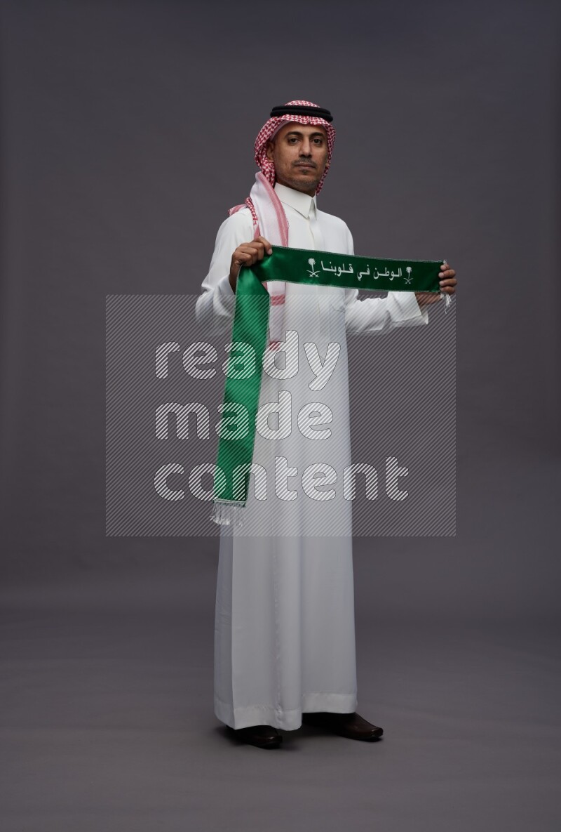 Saudi man wearing thob and shomag standing holding Saudi flag on gray background