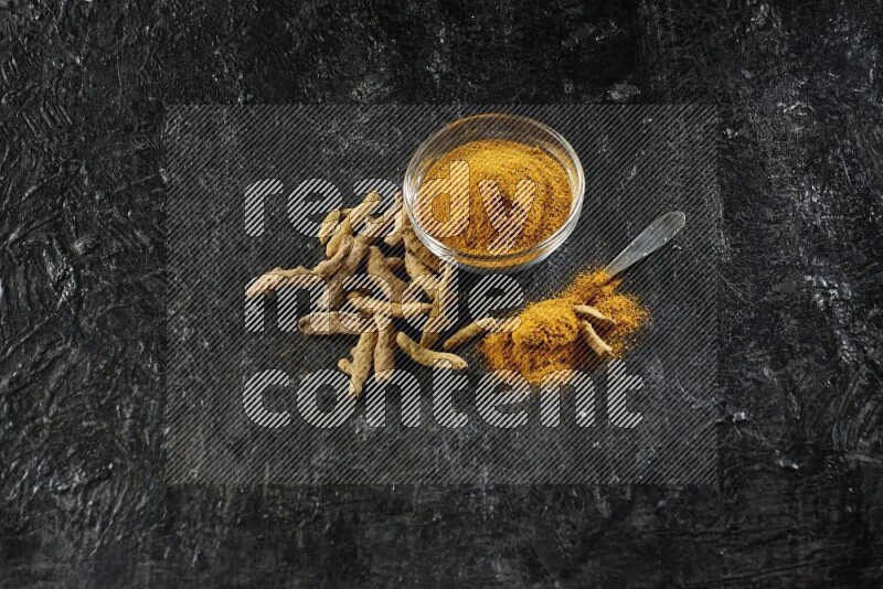 وعاء زجاجي وملعقة معدنية ممتلئان ببودرة الكركم وأصابع الكركم المجففة بجانبهما على خلفية سوداء