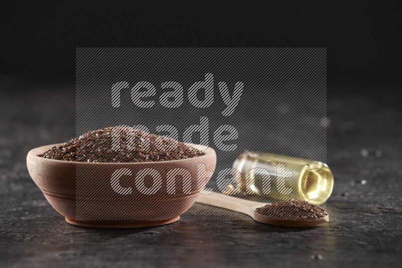 A wooden bowl and spoon full of flaxseeds and a glass bottle of flaxseeds oil on a textured black flooring