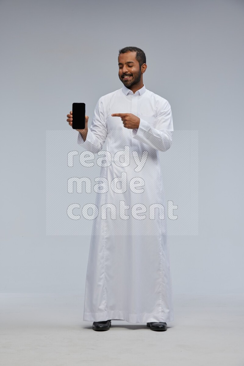 Saudi man wearing Thob standing showing phone to camera on Gray background