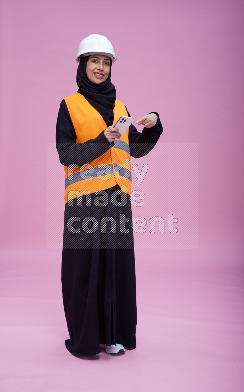 Saudi woman wearing Abaya with engineer vest and helmet standing texting on phone on pink background