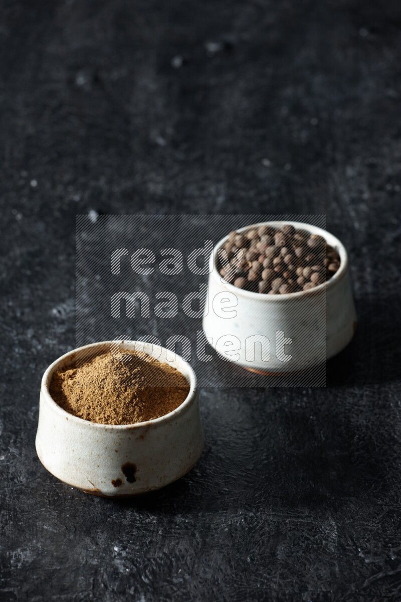 2 beige bowls, one full of allspice powder and the other full of whole balls on a textured black flooring