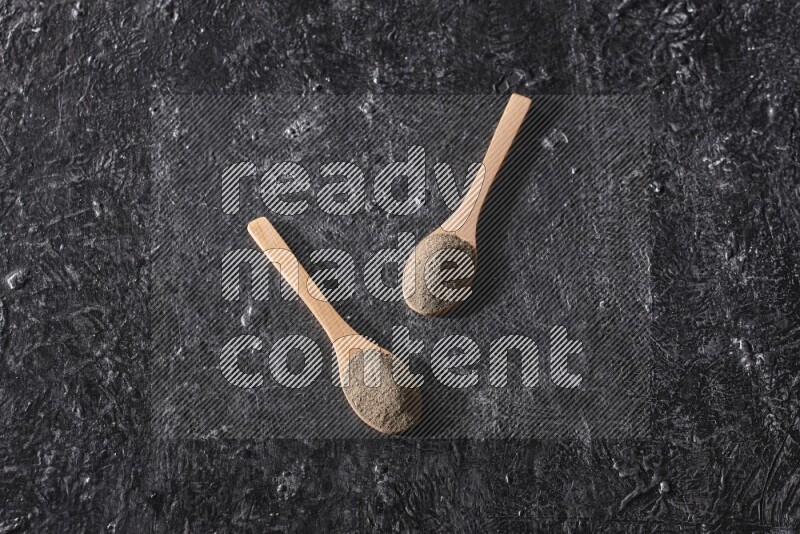 2 wooden spoons full of black pepper powder on a textured black flooring