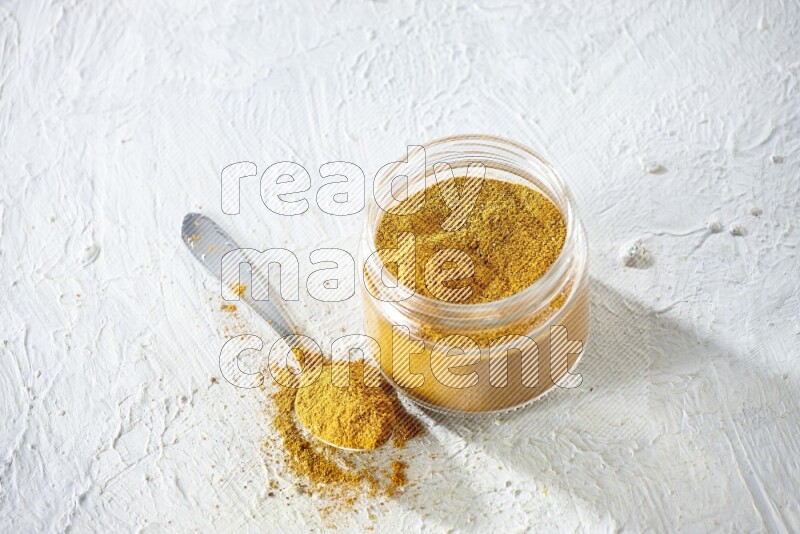 A glass jar and a metal spoon full of turmeric powder on a textured white flooring