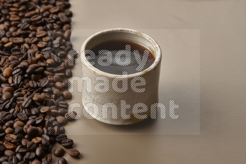 A beige pottery cup of coffee surrounded by roasted coffee beans on beige background
