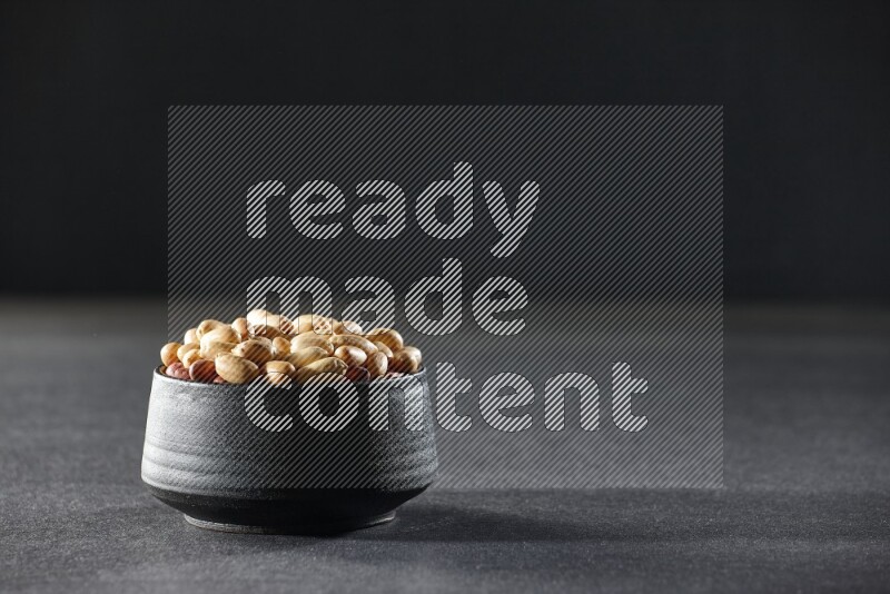 A black pottery bowl full of peeled peanuts on a black background in different angles