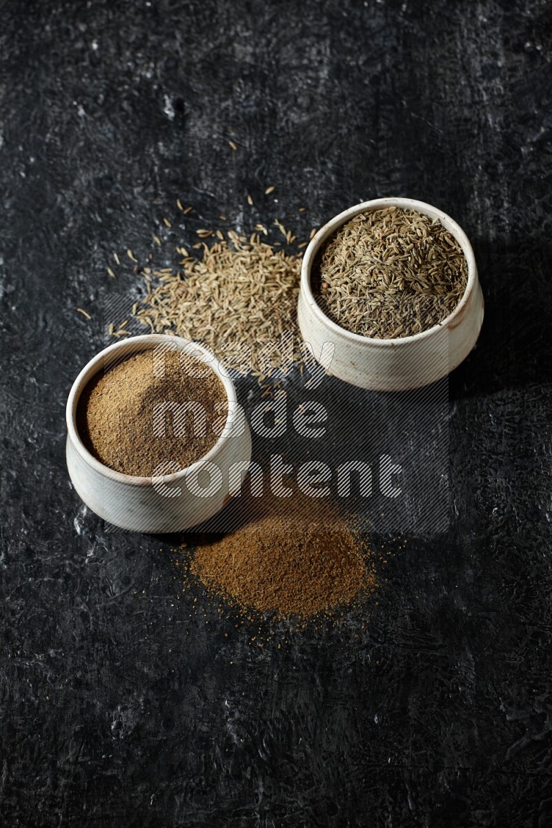 2 beige bowls, one full of cumin powder and the other full of seeds on a textured black flooring