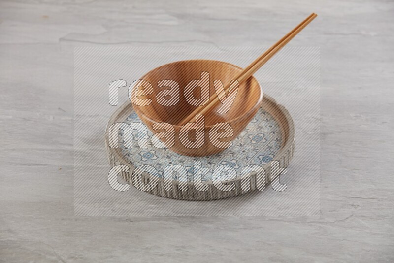 brown wood round bowl on top of multi color round ceramic plate and chopsticks, on grey textured countertop