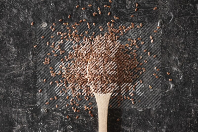 A wooden spoon full of flaxseeds and surrounded by seeds on a textured black flooring