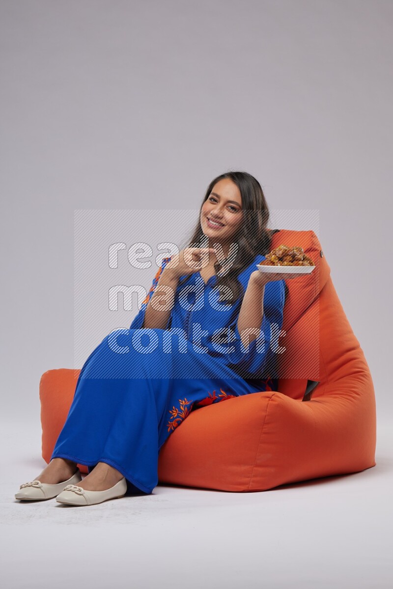 A Woman sitting on an orange beanbag wearing Jalabeya holding a plate of dates