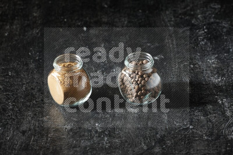 2 glass spice jars full of allspice powder and whole balls on a textured black flooring