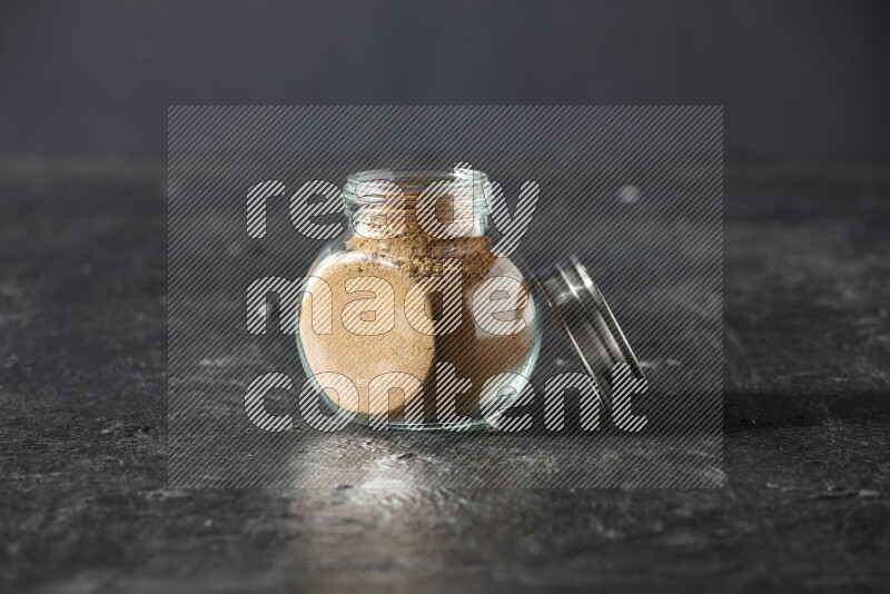 A glass spice jar full of allspice powder on a textured black flooring