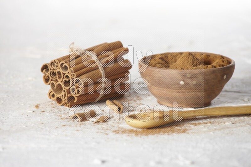 Cinnamon sticks stacked and bounded beside a wooden bowl full of cinnamon powder and a wooden spoon full of powder on white background
