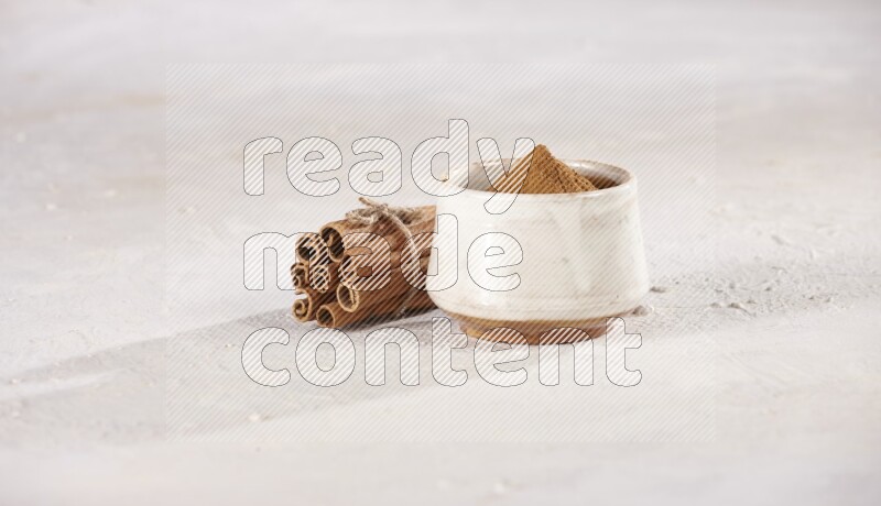 Cinnamon sticks stacked and bounded beside a beige bowl full of cinnamon powder on white background