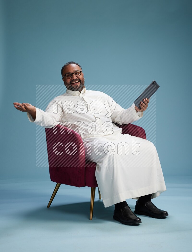 Saudi Man without shimag sitting on chair working on tablet on blue background