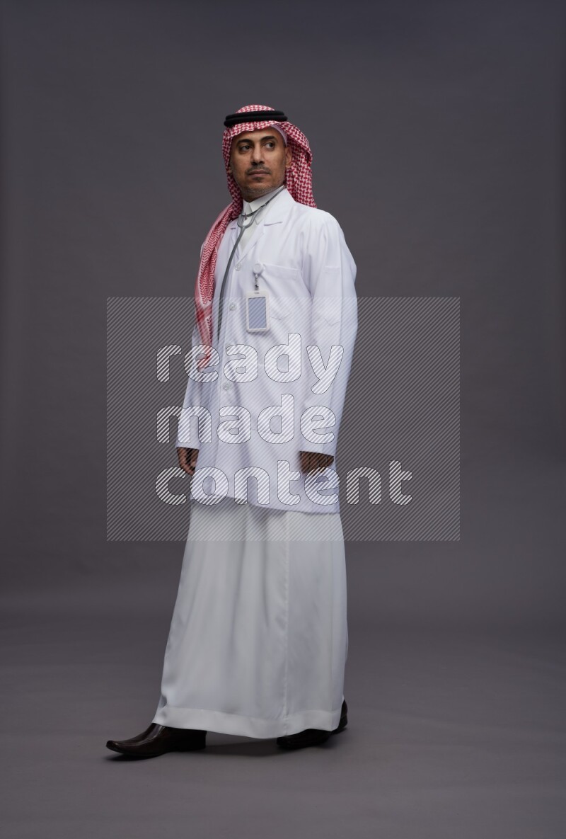 Saudi man wearing thob with lab coat and shomag with pocket employee badge with stethoscope standing interacting with the camera on gray background
